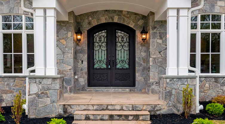 An arts and crafts style front porch with double posts, flagstone stoop, stone wall, arched doorway and coach style lights.