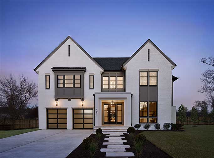 A front home exterior blending Modern Tudor and Farmhouse, with patterned panel and window assemblies, white painted brick, aluminum garage door with smoked glass and custom details.