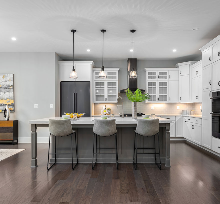 A modern farmhouse kitchen with subway tile backsplash, quartz countertops, grey center island base and white cabinets, some with glass doors. Dining area off to the left of the photo.