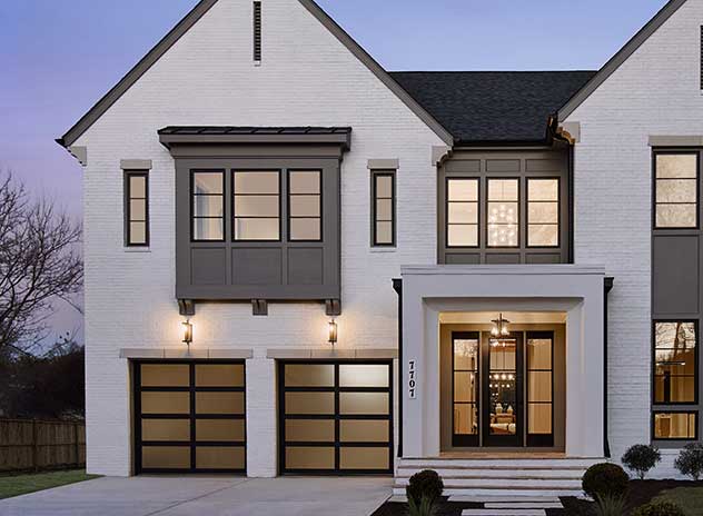 A front home exterior blending Modern Tudor and Farmhouse, with patterned panel and window assemblies, white painted brick, aluminum garage door with smoked glass and custom details.