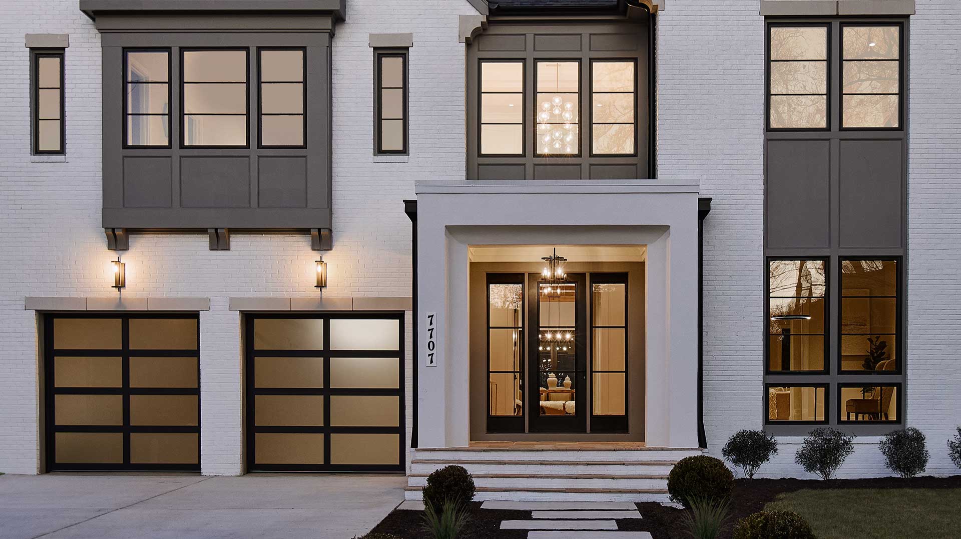 A close up of the front exterior of a home with patterned panel and window assemblies, white painted brick, aluminum garage door with smoked glass and custom details.