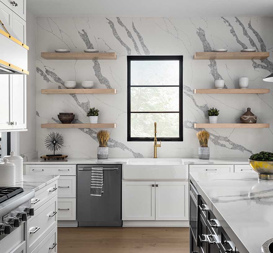 An accent wall of Q Premium Natural Quartz with floating natural wood shelves, farmhouse-style sink, and matching countertop. Quartz kitchen island and professional cooktop in the foreground.