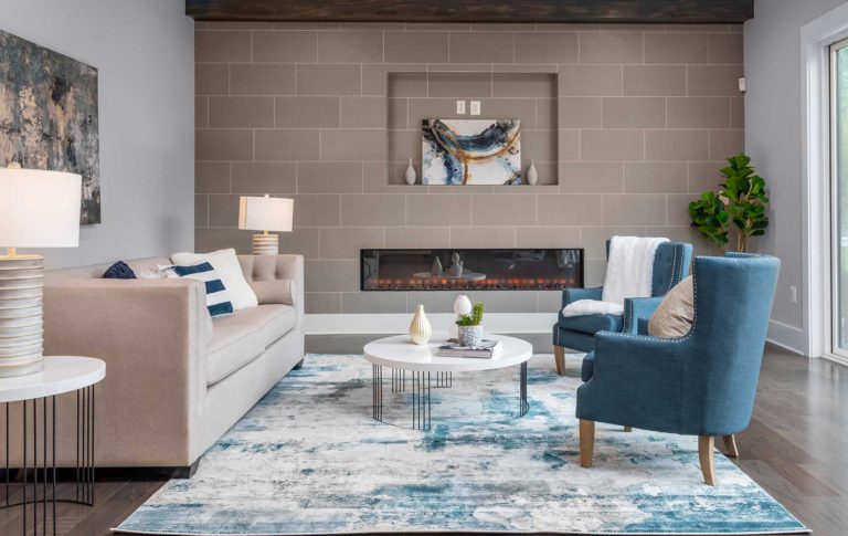 A modern farmhouse style family room with oversized tile accent wall with linear fireplace and TV niche, exposed natural stained beams. Blue and neutral furniture in the foreground.