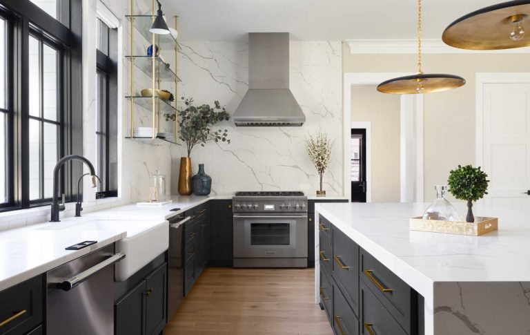 A kitchen with farmhouse sink and Brizo Litze® fixture in matte black and luxe gold, quartz watefall island with dark cabinets beneath, range and fume hood at far end