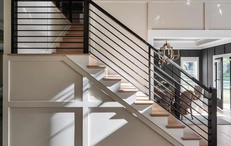 The side of a staircase with paneled walls, horizontal black iron rails, white risers with natural wood stair treads