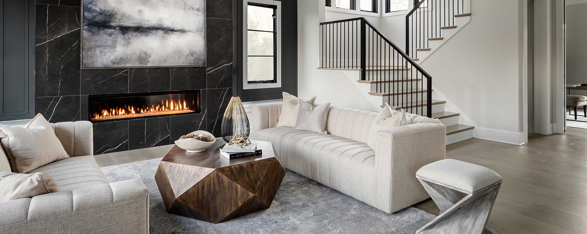 An oversized linear fireplace surround of dark Daltile Marble Attaché with dark windows and walls, stairs with horizontal iron rails in the background, neutral color furniture in the foreground