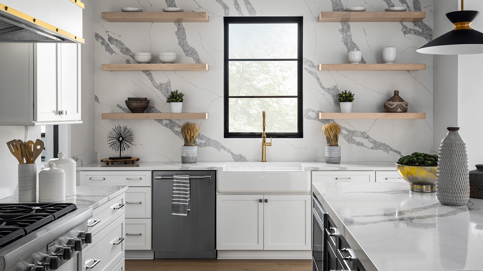 An accent wall of Q Premium Natural Quartz with floating natural wood shelves, farmhouse-style sink, and matching countertop. Quartz kitchen island and professional cooktop in the foreground.