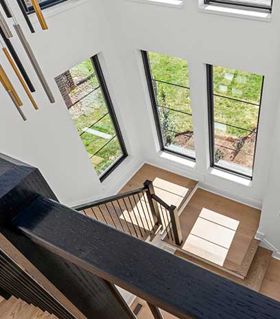 Looking down from the top of a stair case lined with tall black windows, dark stained railing and contemporary chandelier