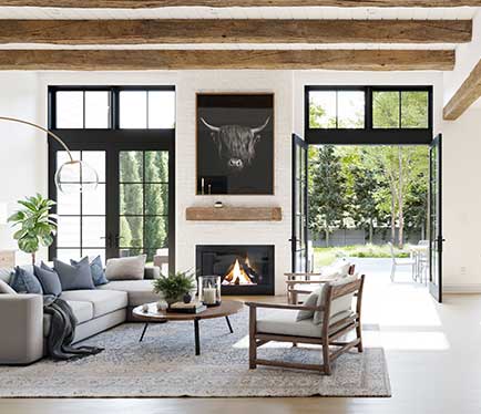 A modern farmhouse style family room with stained exposed beams, fireplace with natural wood hearth and a pair of double exterior doors.