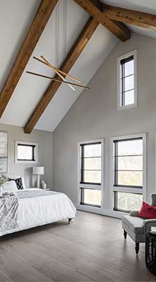A modern farmhouse style master bedroom with stained exposed beams, high vaulted ceiling, and floor height dark frame windows.