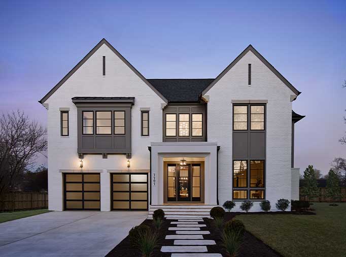 A front home exterior blending Modern Tudor and Farmhouse, with patterned panel and window assemblies, white painted brick, aluminum garage door with smoked glass and custom details.