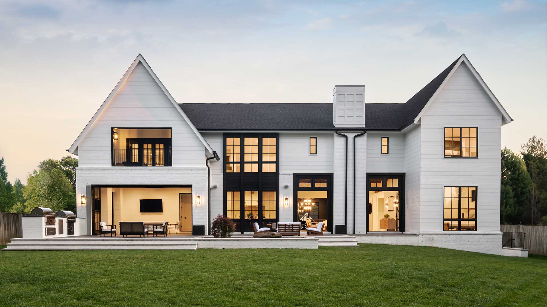The rear of a custom modern farmhouse with an indoor outdoor space open the terraced rear patio, additional exterior doors also open, with lights in all windows against a darkening sky.