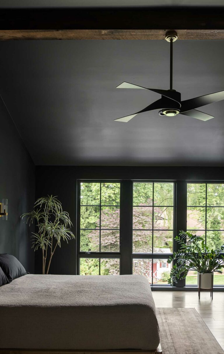 A modern farmhouse owner's suite bedroom with dark walls and dark windows, with a bright green backyard visible outside, modern ceiling fan above