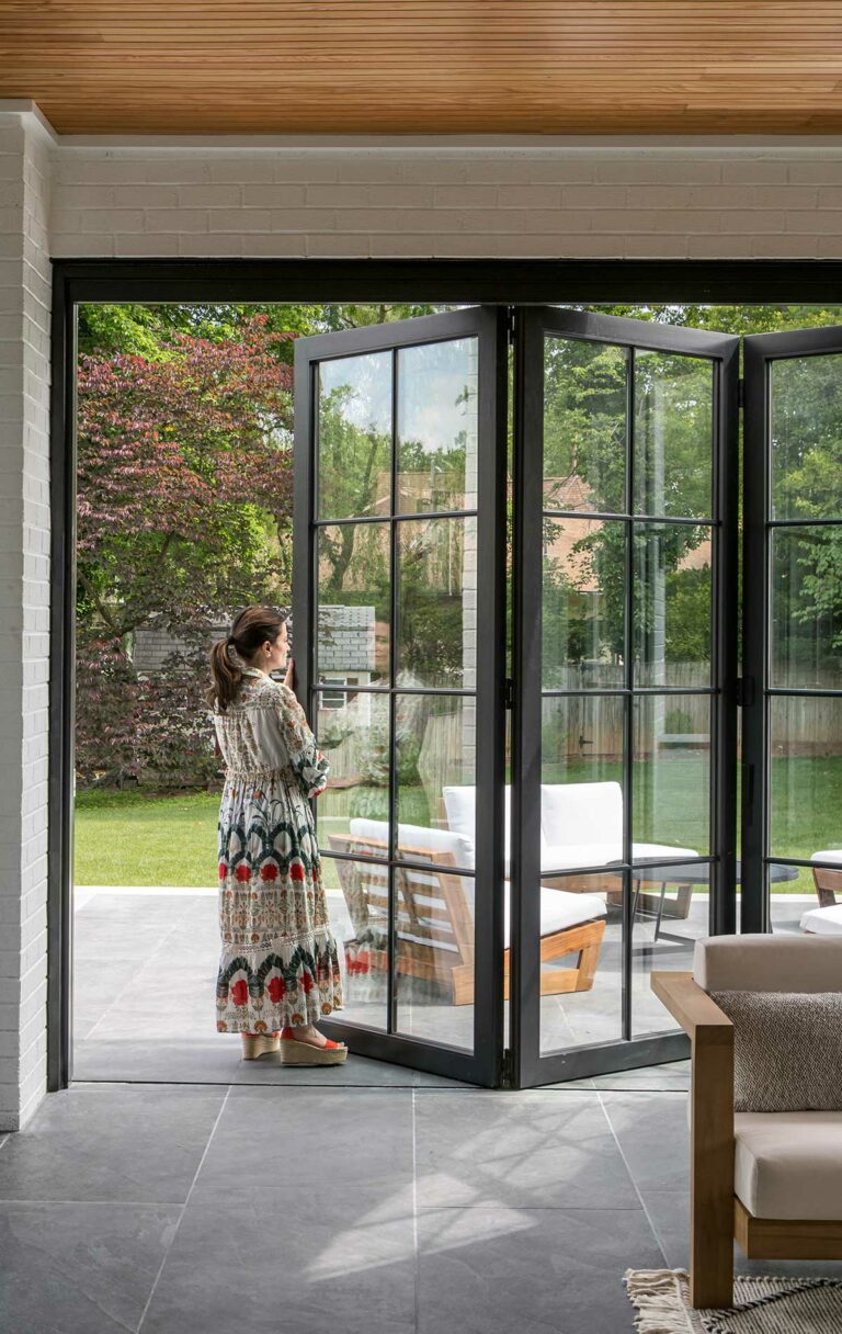 A woman stands at a folding patio door, opening to a patio and green backyard.