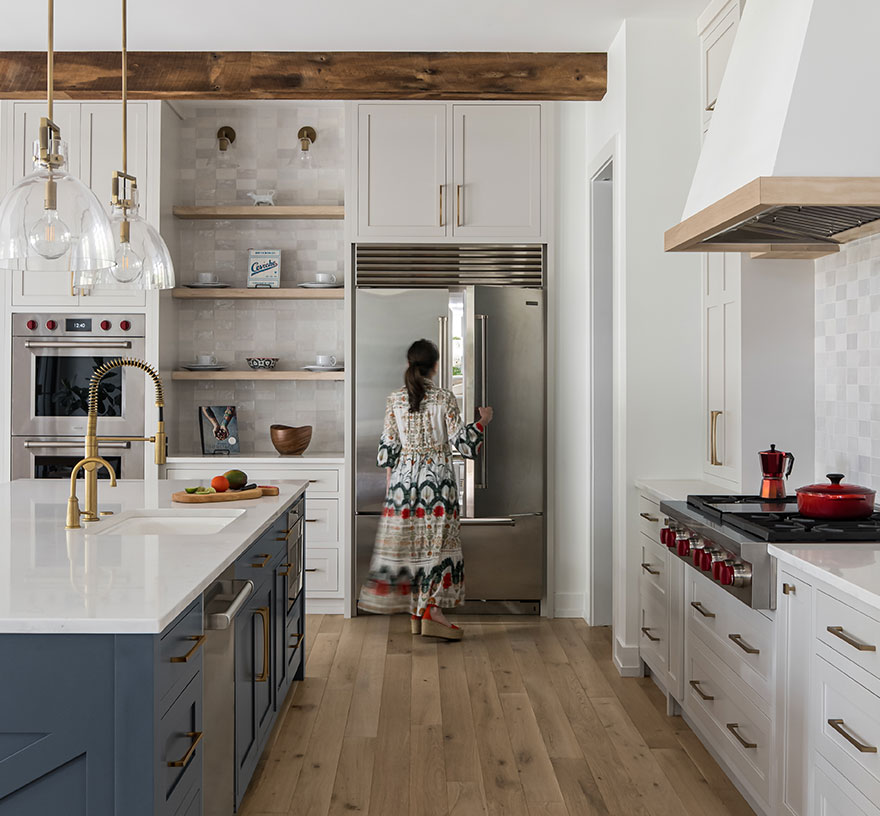 A woman walks toward the refrigerator from the far side of a modern farmhouse kitchen with blue island, exposed beams, tile backsplash and accent wall