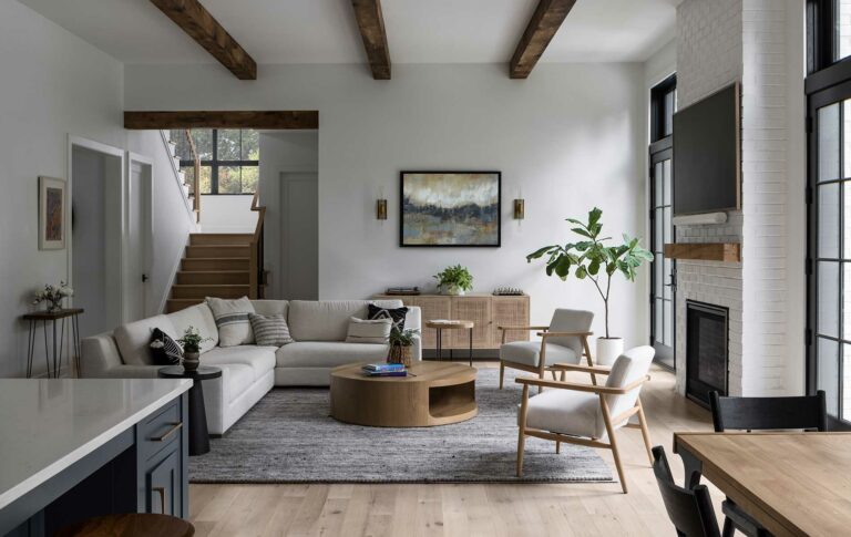A modern farmhouse living room with exposed natural wood stained beams and matching fireplace mantel. Staircase in the background.