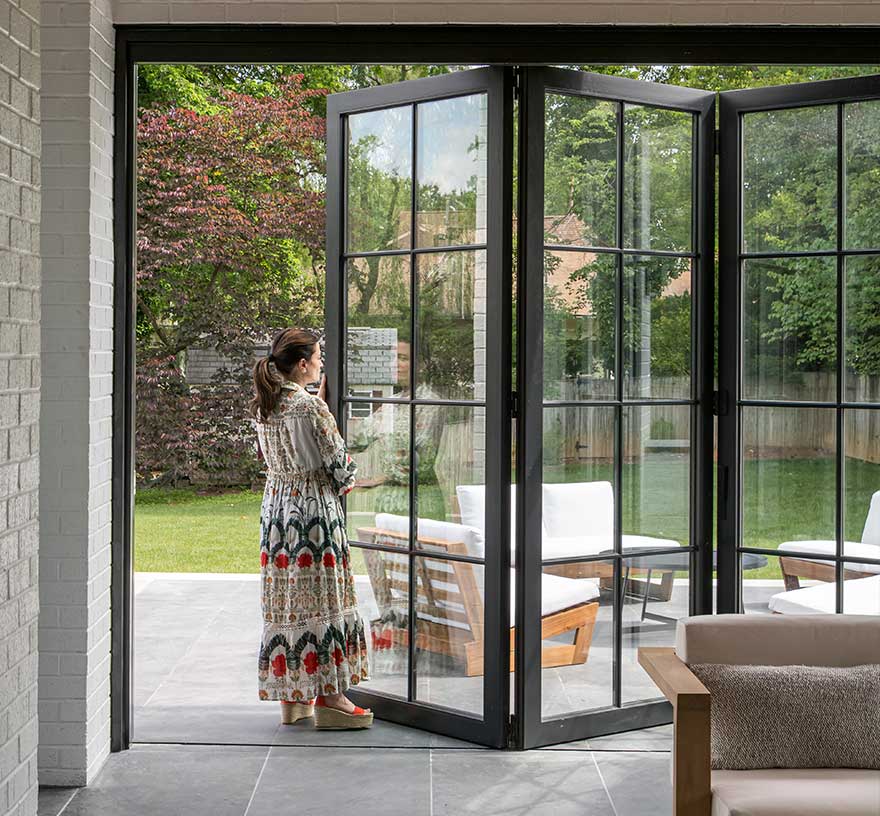 A woman stands at a folding patio door, opening to a patio and green backyard.