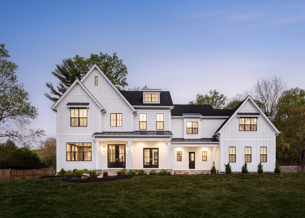 Twilight photo of a lit up modern farmhouse front exterior in white board and batten with two covered entrances and dark colored windows.