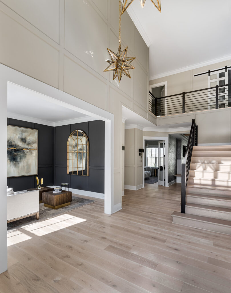 A tall photo showing a modern two-story foyer, dark paneled living room to the left, stairs to the right, white risers, natural stairs, iron rails