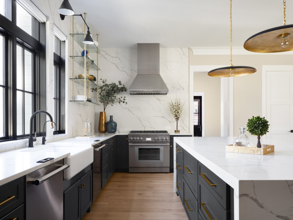 A kitchen with farmhouse sink and Brizo Litze® fixture in matte black and luxe gold, quartz watefall island with dark cabinets beneath, range and fume hood at far end