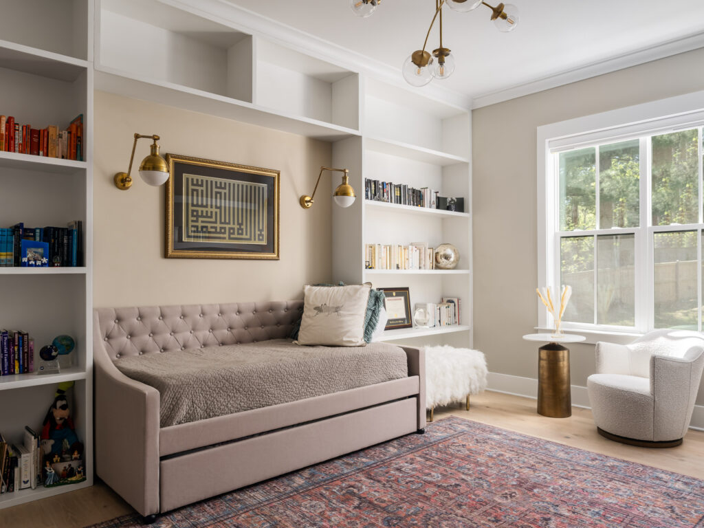 A daybed against a wall of built in white shelving filled with books, a small table and chairs sit in front of a window to the right.