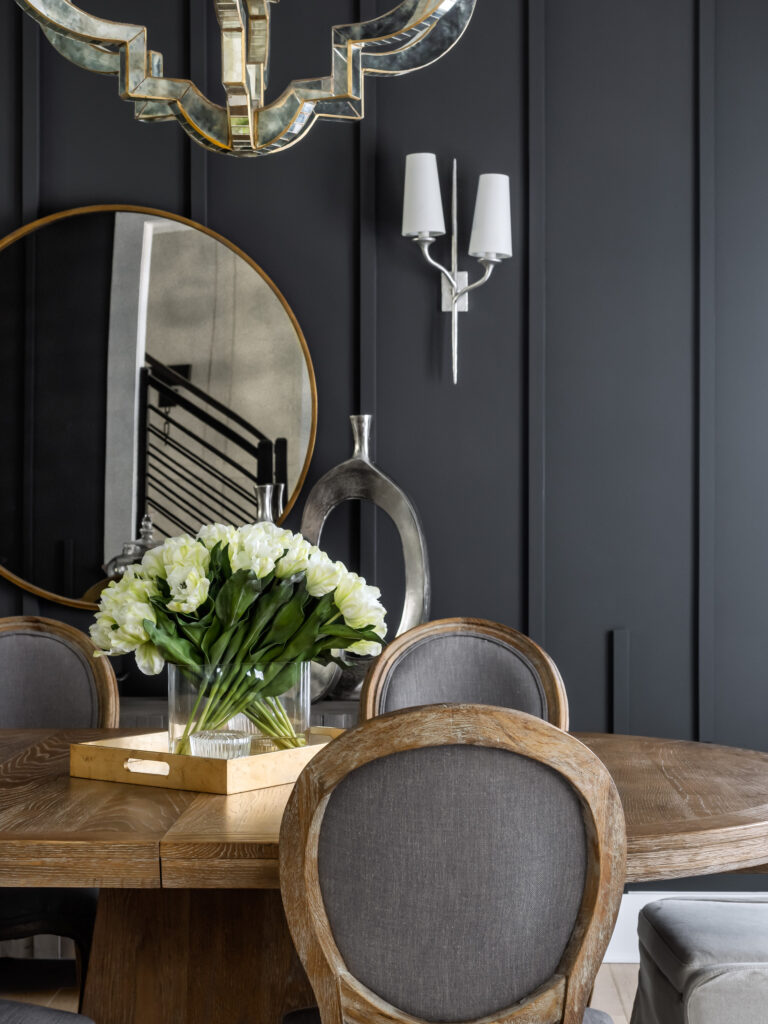 A detail of a dining room with dark paneled accent wall and modern mirrored chandelier, stairs visible in a round mirror on the wall, table and chairs in the foreground.