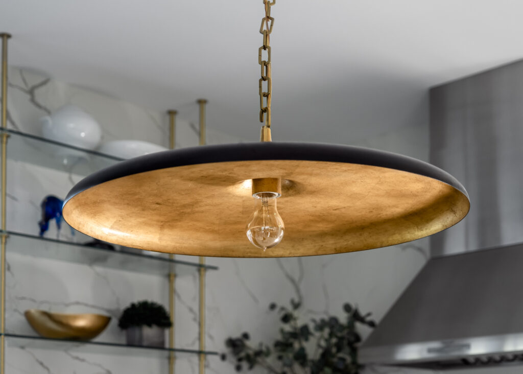 A detail image of a hanging kitchen light fixture, gold on the underside with exposed bulb, black on top with a gold chain.