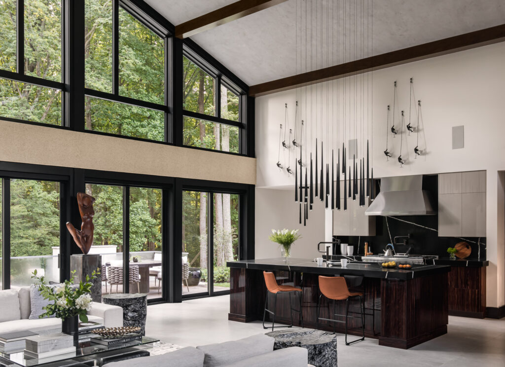 A gabled glass wall showing a family room in the foreground with a gourmet Kitchen with contemporary lighting, commercial depth range, frameless cabinets and Silestone Eternal Noir countertops and backsplash to the right.