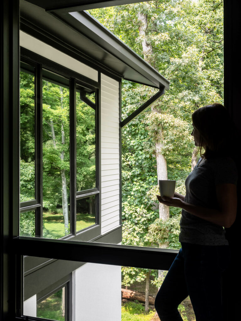 A woman looks out a large picture window at the reflection of the forest in another set of picture windows.