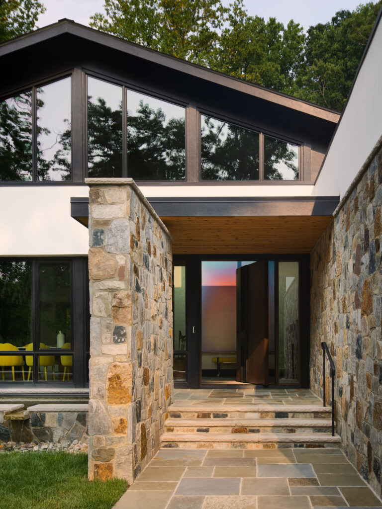 The front entrance to the home, a stone wall to the left, pivoting door ahead with trees reflecting in the windows in the gable above.