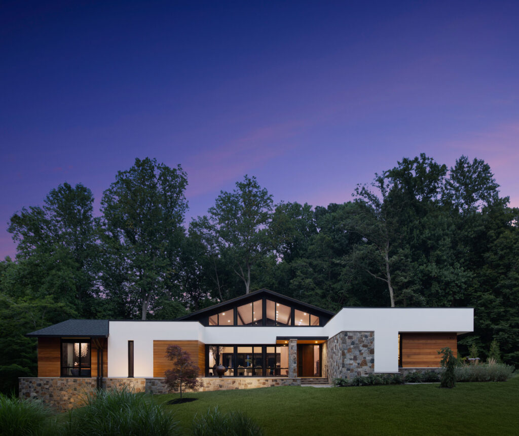 A California Modern style home of stucco, stained natural wood planks, stone and tall custom modern windows against tall trees and a twilight sky.