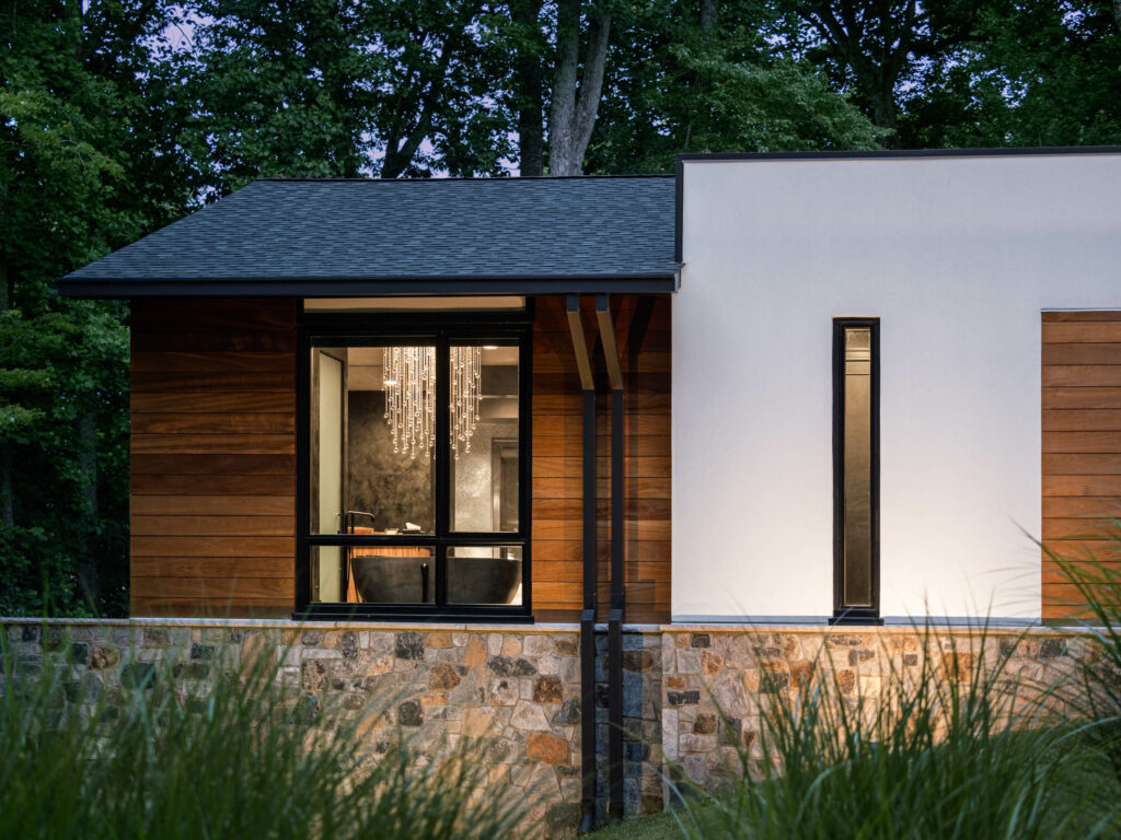 An Owner's Bath and chandelier glow through windows at dusk on a wall of stained wood panels beside stucco