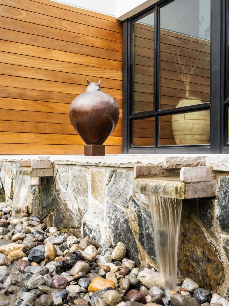 A fountain drains water from the reflecting pool above the stone wall into a bed of river rock, owl sculpture visible above.