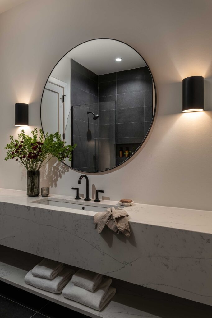 A contemporary vanity in grey quartz shows a black tile shower reflected in a circular mirror.