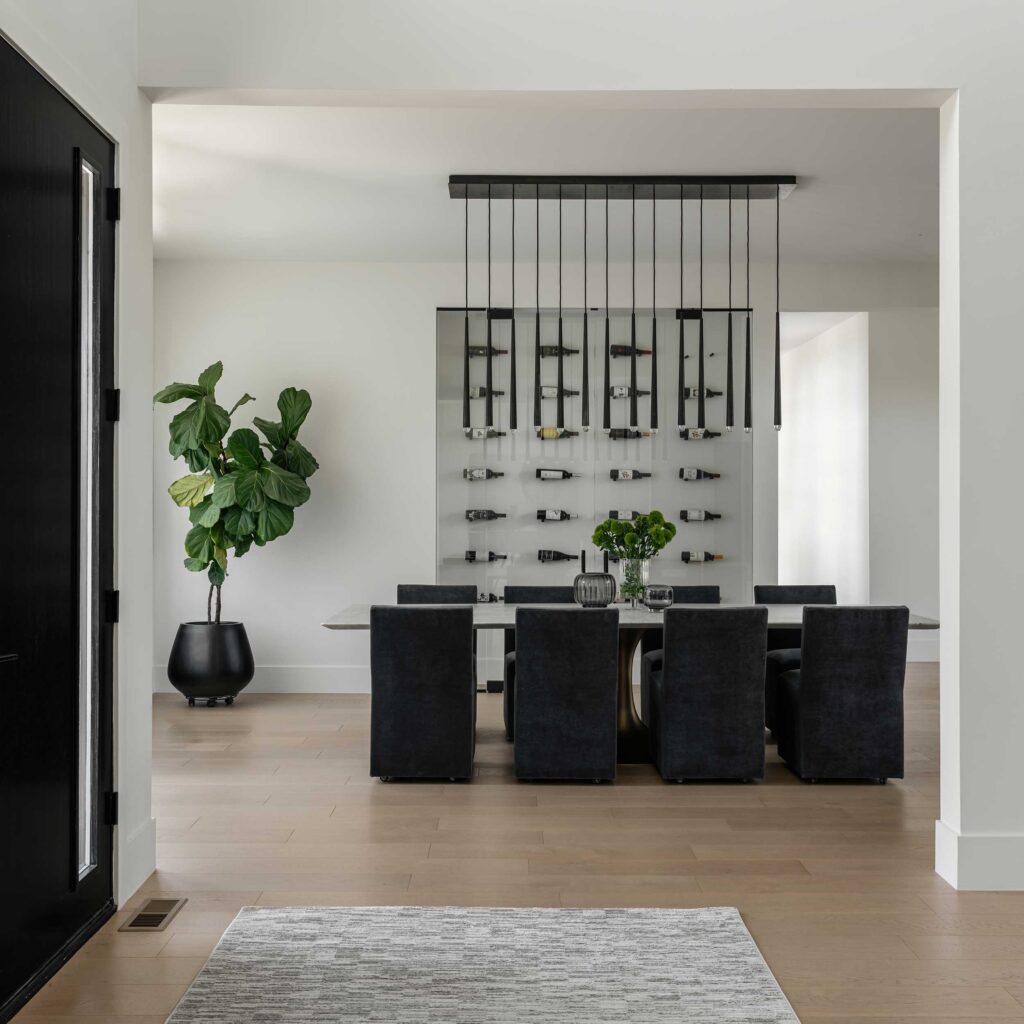 Looking into a dining room from the foyer, a dark chandelier and dark chairs contrast against a white room with light wood floors and wall display of horizontal wine bottles behind glass doors.