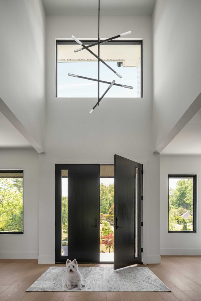 A two story foyer with modern chandelier, large dark grill-less windows, dark double front door with sidelights. A west highland terrier sits on a rug in the foreground.