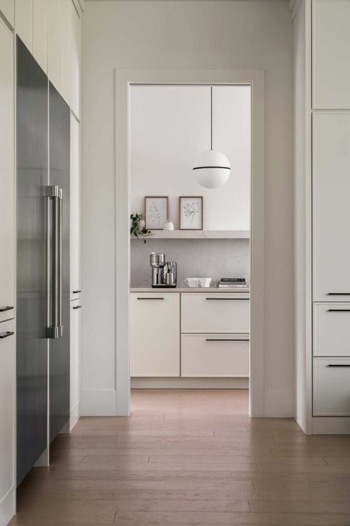 A detail photo looking through a door into the Butler's Pantry with white cabinetry and white globe light hanging from the ceiling. A large stainless steel refrigerator is to the left in the foreground.