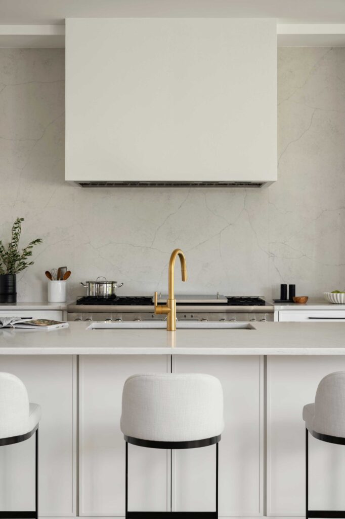 A kitchen detail photo showing a white island with white quartz top and brass fixture, behind an oversized stainless steel range against a white quartz backsplash.