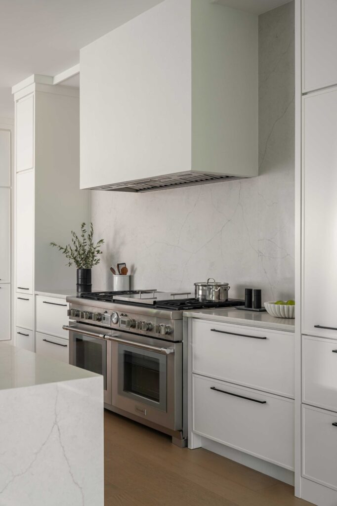 A kitchen detail showing a large professional stainless steel range, fume hood covered in painted white, quartz backsplash and counters, white cabinets.