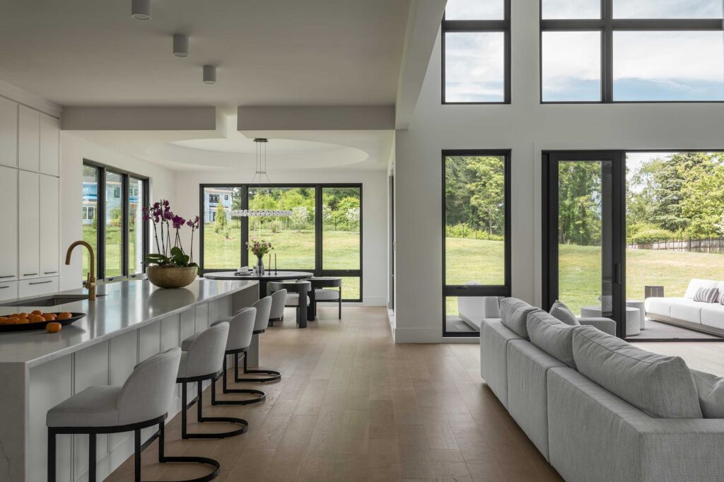A wide photo showing a contemporary white kitchen to the left, morning room beyond the kitchen with large windows and circular tray ceiling, and family room to the right, with large windows and patio doors open to the outside.