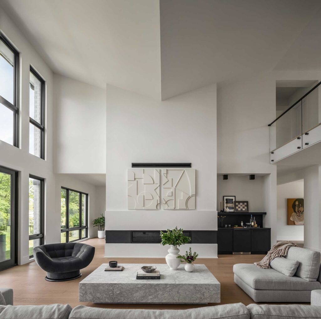 A two story family room with a vaulted butterfly ceiling, second floor overlook, linear fireplace and dry bar. Contemporary art hangs over the fireplace, tall windows to the left.