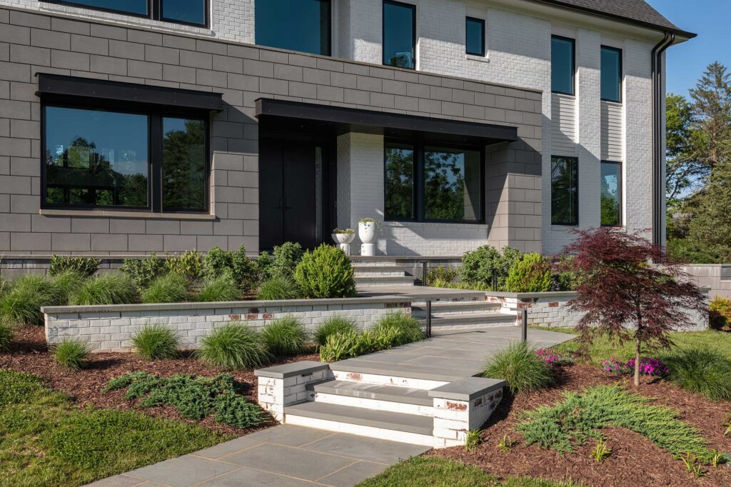 A photo of a flagstone leadwalk with powdered white brick risers and garden walls. The home entrance is surrounded by a large geometric shape of grey stone block, the house is white brick with grill-less windows.