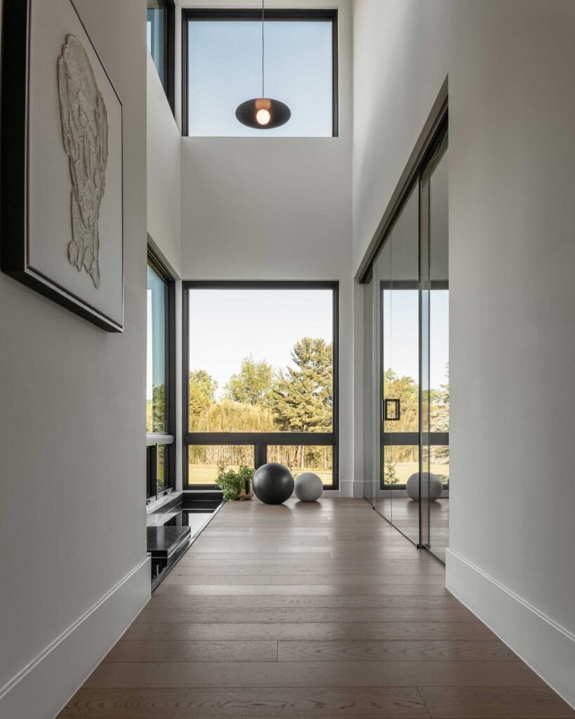 A two story space with oversized dark framed windows, a narrow reflecting pool to the left and glass doors to the right. Modern artwork hangs in the foreground.