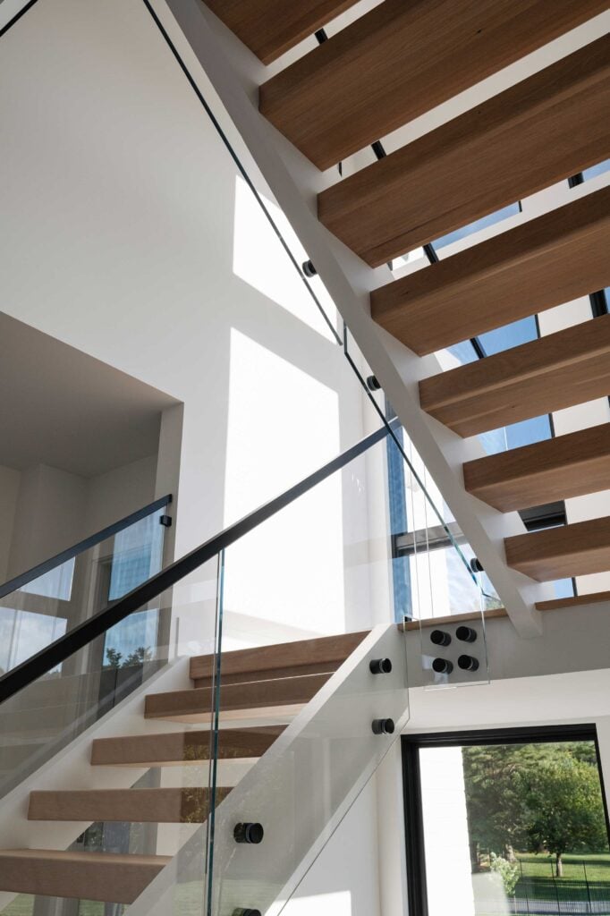 A close up image of an open riser staircase with glass walls, black handrails and stained steps. Windows line the entire staircase.