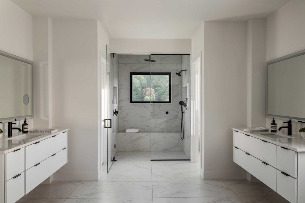 An owner's suite bath with dual floating vanities on either side in the foreground, white cabinetry and counterops, black fixtures. Beyond a glass walled shower with wall and overhead showerheads and quartz wall and seat.