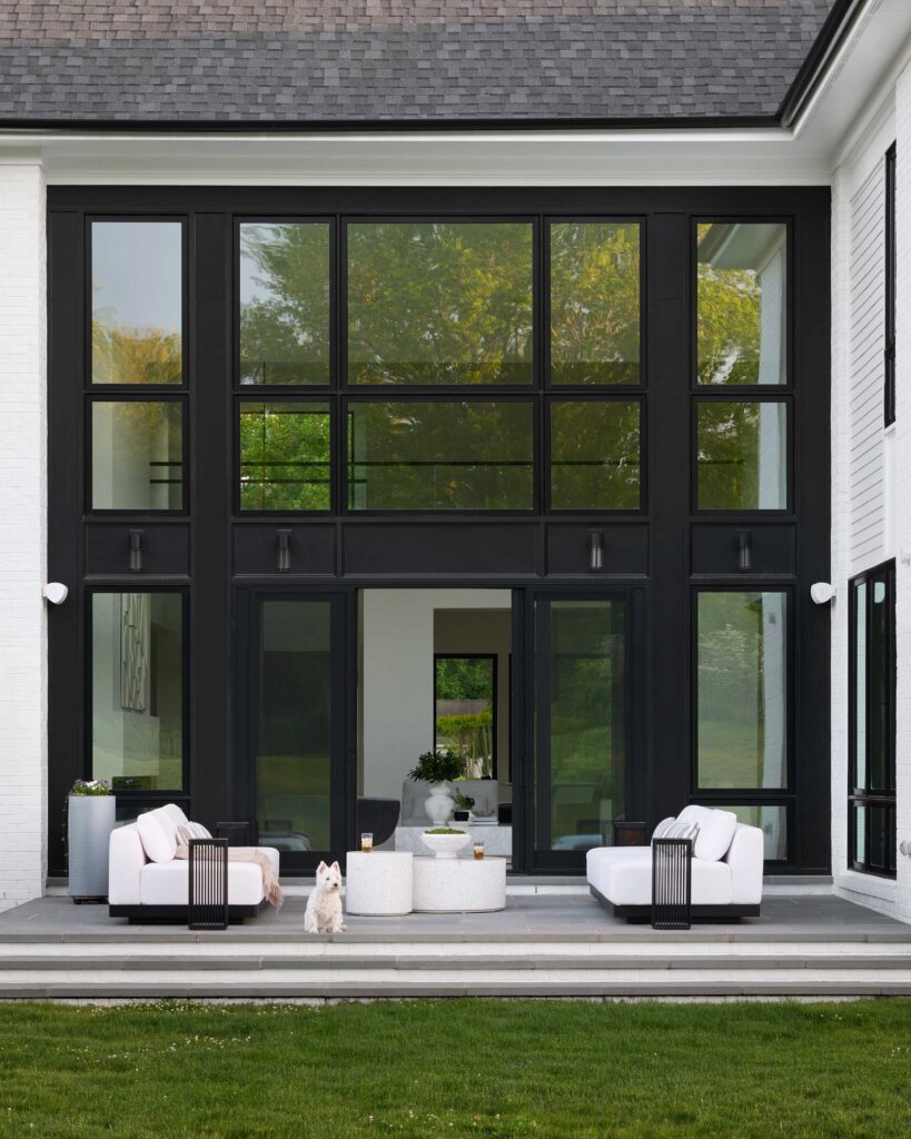 A rear patio photo, white couches and tables, dark side tables and dark frame oversized windows with sliding doors open to inside.