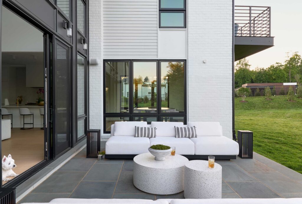 A patio with white round tables, white couch and dark side tables. A large sliding door is open to the Family Room with the Kitchen visible, a dog sits in the doorway. A balcony is visible above.