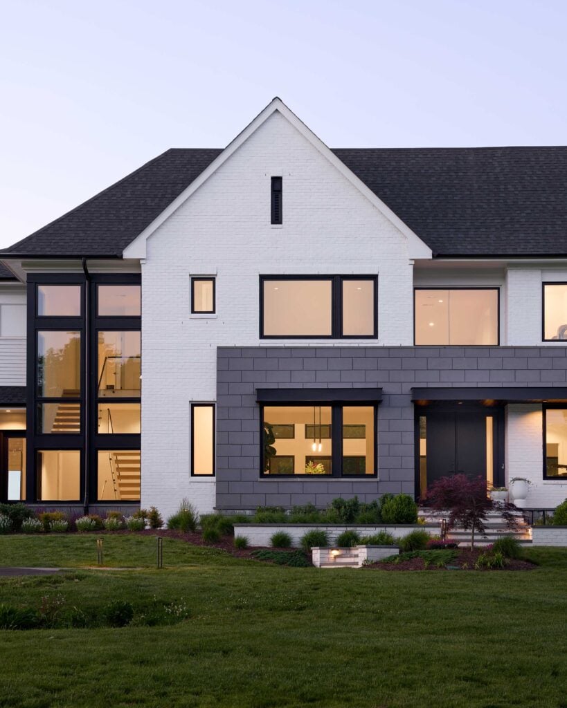 A detail photo showing the front of a home in the evening, both stories of a stair case clearly visible through tall black framed modern windows. Grey block surrounds the front door and large 1st floor window.