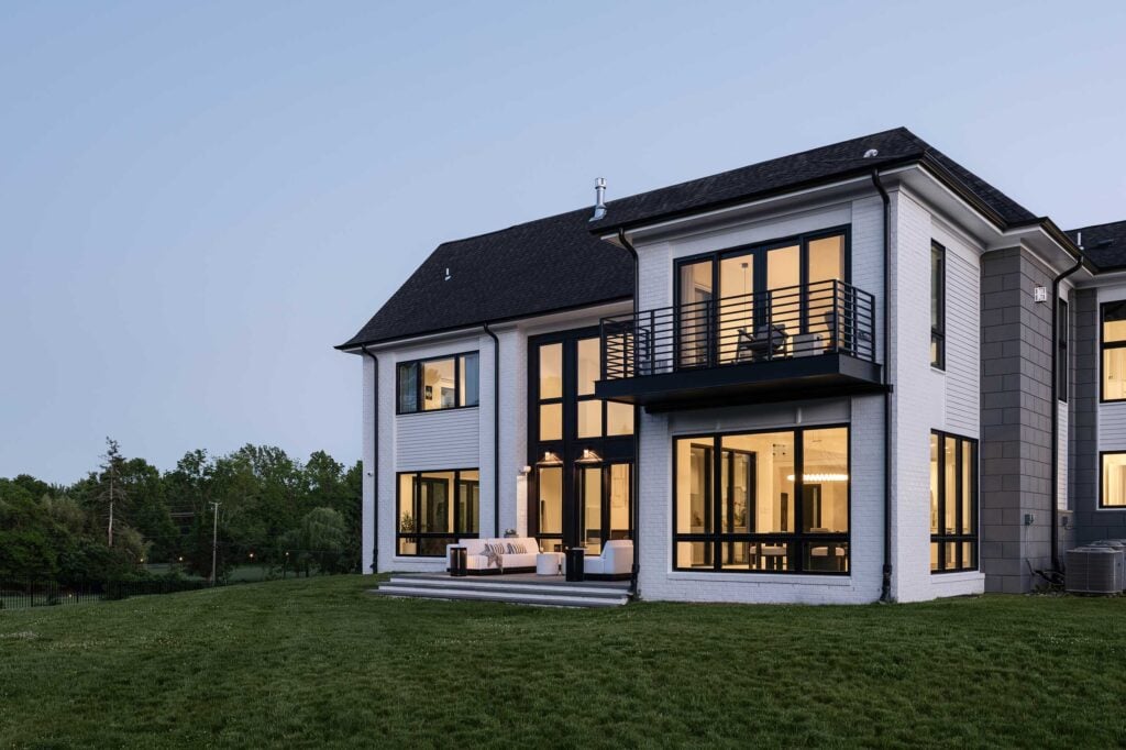 The rear exterior of a home lit up in the early evening, a balcony off a second floor bedroom, tall windows across the house, couch and tables on the patio, grey block accents.