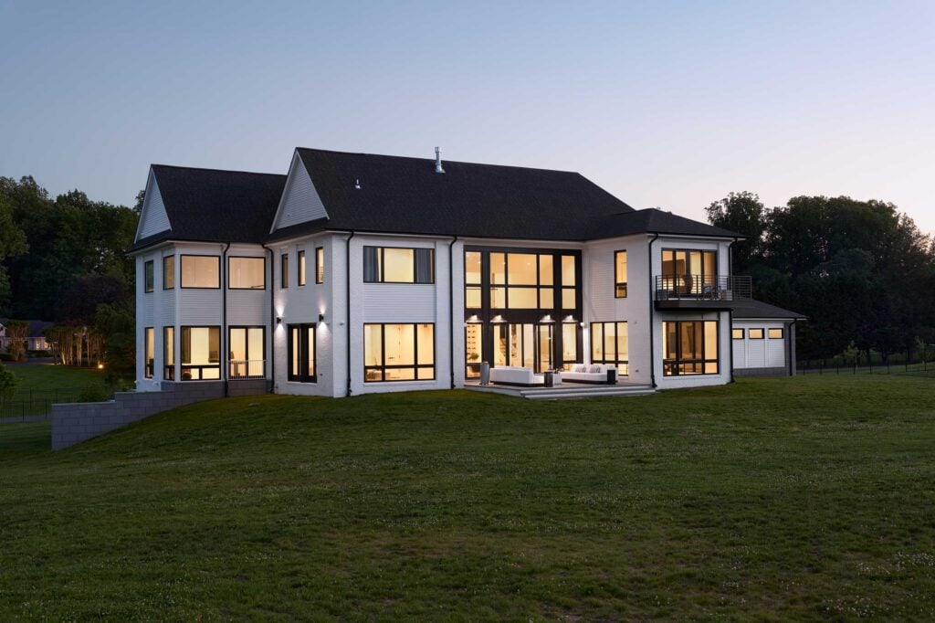 The rear exterior of a home lit up in the early evening, a balcony off a second floor bedroom, tall windows across the house, couch and tables.